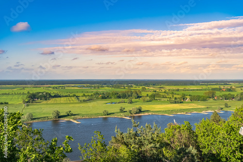 View over the Elbauen in Lower Saxony, Germany. You see a landscape with fields, meadows and the river Elbe under a blue sky with clouds, which are illuminated by the sun.