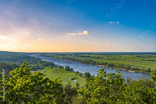 View over the Elbauen in Lower Saxony, Germany. You see a landscape with fields, meadows and the river Elbe under a blue sky with clouds, which are illuminated by the sun.