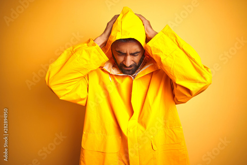 Young indian man wearing raincoat with hood standing over isolated yellow background suffering from headache desperate and stressed because pain and migraine. Hands on head. © Krakenimages.com