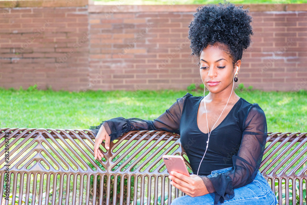 Young beautiful African American Woman with afro hairstyle wearing mesh sheer long sleeve shirt blouse, sitting on bench at park in New York, listening music with earphone and cell phone, reading..