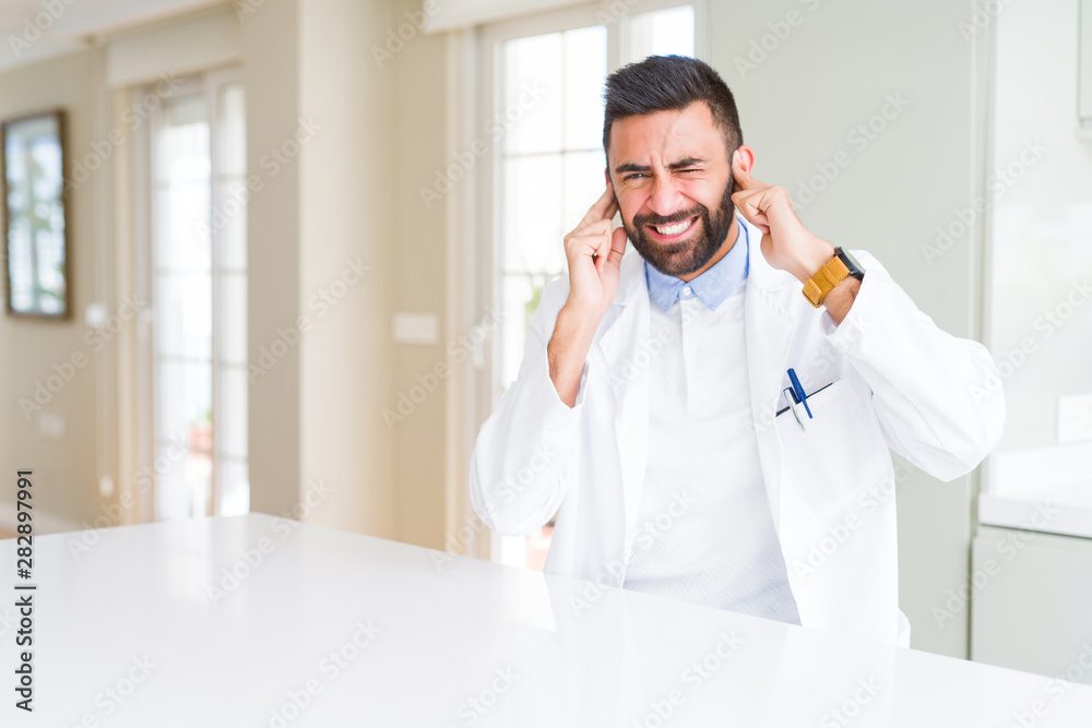 Handsome hispanic doctor or therapist man wearing medical coat at the clinic covering ears with fingers with annoyed expression for the noise of loud music. Deaf concept.