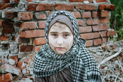 a refugee child in the war, a Muslim girl with a dirty face on the ruins, the concept of peace and war, the child is crying and waiting for help. photo