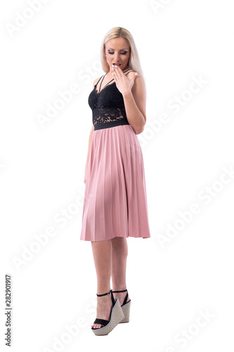 Oops. Shocked young blonde woman with surprised expression covering mouth looking down. Full body isolated on white background. 