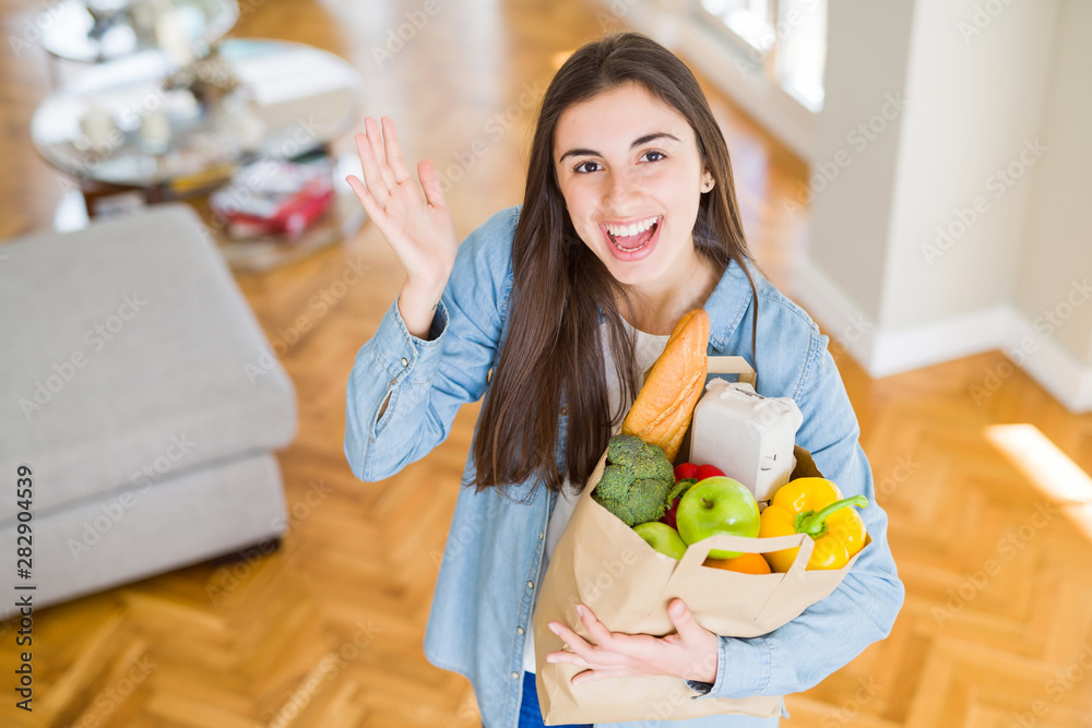 Beautiful young woman holding paper bag full of healthy groceries very happy and excited, winner expression celebrating victory screaming with big smile and raised hands Beautiful young woman holding paper bag full of healthy groceries very happy and excited, winner expression celebrating victory screaming with big smile and raised hands
