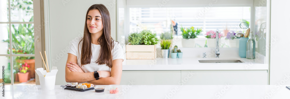 Wide angle picture of beautiful young woman eating asian sushi from delivery smiling looking side and staring away thinking.