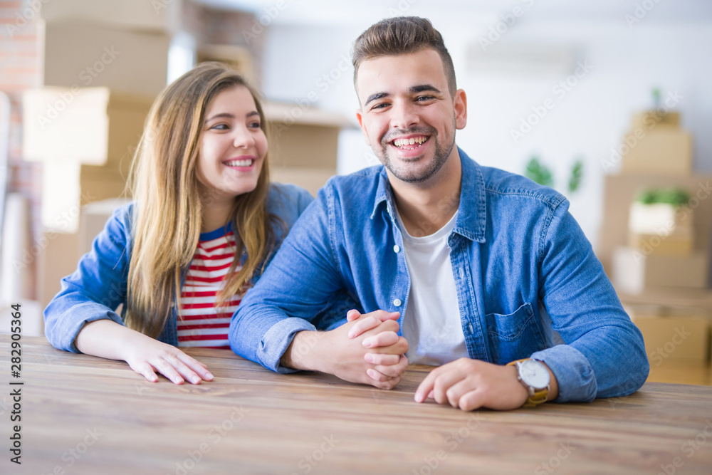 Young beautiful couple sitting on the table at home, hugging in love very happy for moving to new home with cardboard boxes behind them Young beautiful couple sitting on the table at home, hugging in love very happy for moving to new home with cardboard boxes behind them