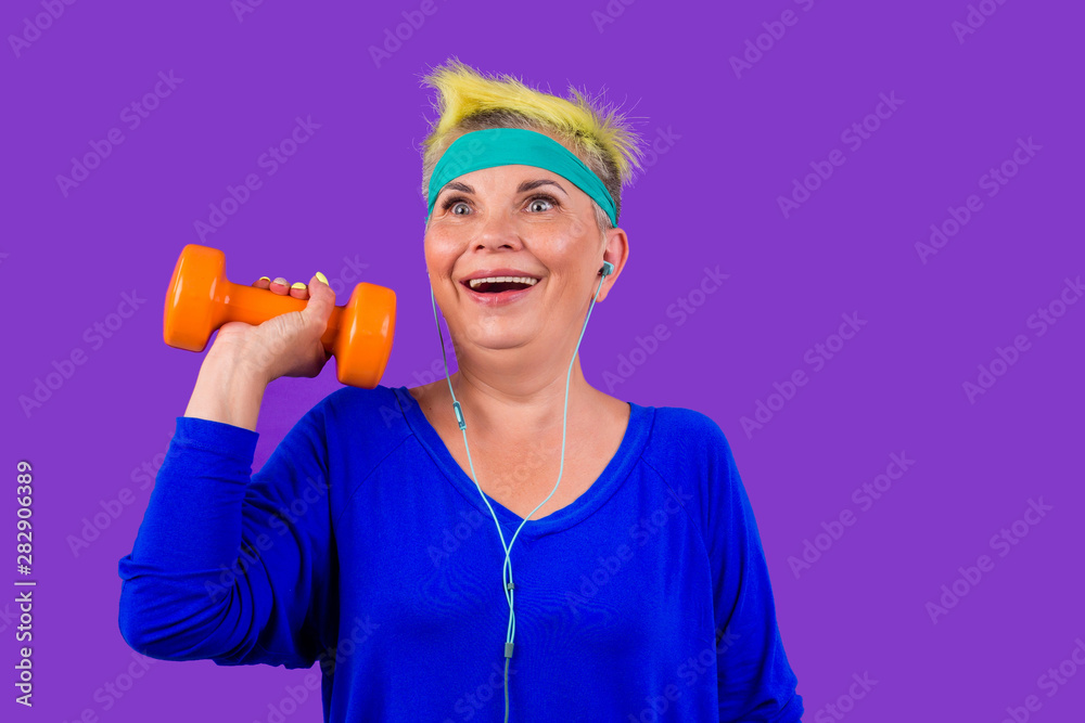 happy active pensioner woman with stylish yellow dyed hair and a blue bandage on forehead after pilates training smiling in studio violet background.forever young soul