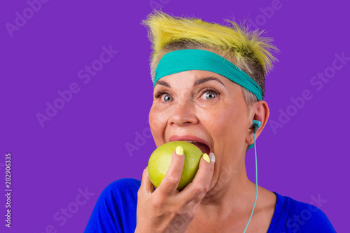 old caucasian woman with short yellow dyed hair and a blue bandage on forehead eating green fresh apple in studio violet background.sporty lady listening to music on headphones