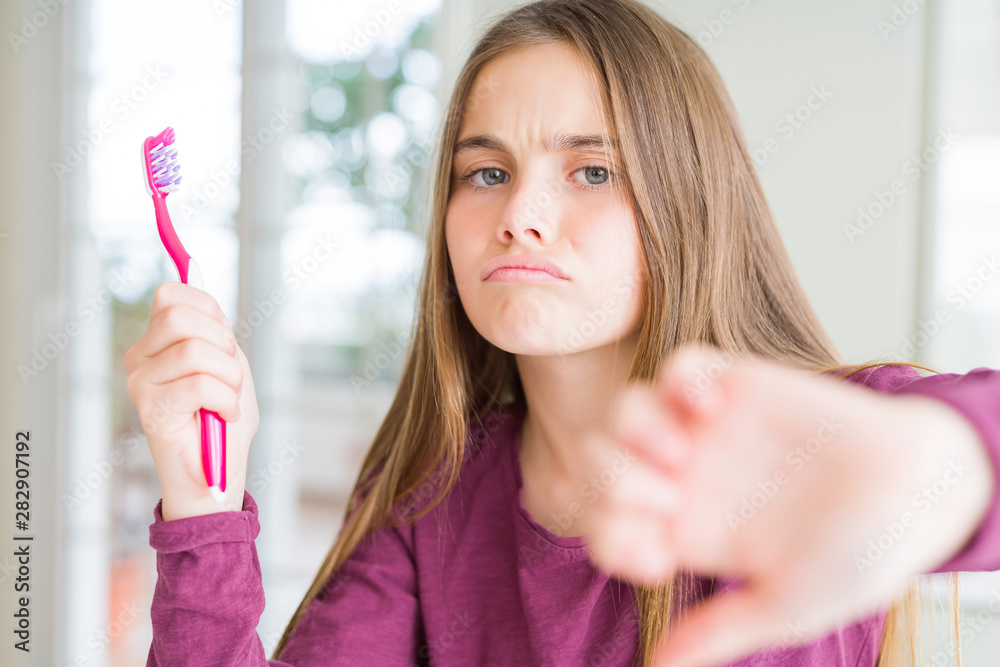 Beautiful young girl kid holding pink dental toothbrush with angry face, negative sign showing dislike with thumbs down, rejection concept Beautiful young girl kid holding pink dental toothbrush with angry face, negative sign showing dislike with thumbs down, rejection concept