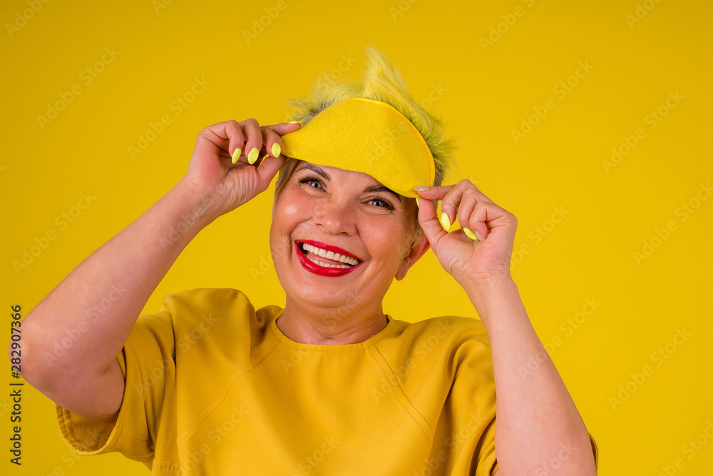 mature woman with colored stylish yellow dyed hair in warm dress with sleeping mask on her eyes background studio copy spase