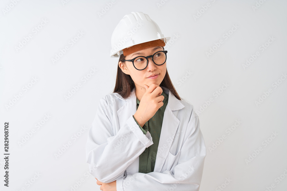 Young chinese engineer woman wearing coat helmet glasses over isolated white background with hand on chin thinking about question, pensive expression. Smiling with thoughtful face. Doubt concept. Young chinese engineer woman wearing coat helmet glasses over isolated white background with hand on chin thinking about question, pensive expression. Smiling with thoughtful face. Doubt concept.