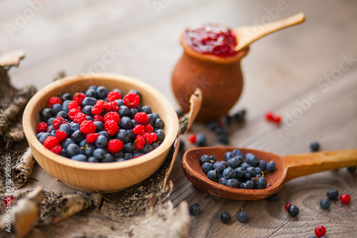 Blueberry and raspberry berry jam in a clay pot, a wooden spoon with fresh berries and birch bark on a wooden background
