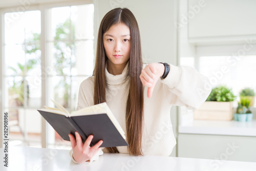 Beautiful Asian woman reading a book with angry face, negative sign showing dislike with thumbs down, rejection concept