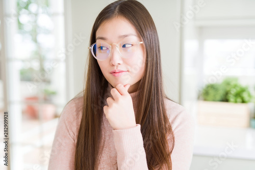 Beautiful Asian woman wearing glasses with hand on chin thinking about question, pensive expression. Smiling with thoughtful face. Doubt concept.