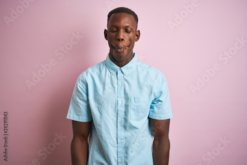 African american man wearing blue casual shirt standing over isolated pink background making fish face with lips, crazy and comical gesture. Funny expression.