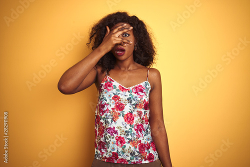 African american woman wearing floral summer t-shirt over isolated yellow background peeking in shock covering face and eyes with hand  looking through fingers with embarrassed expression.