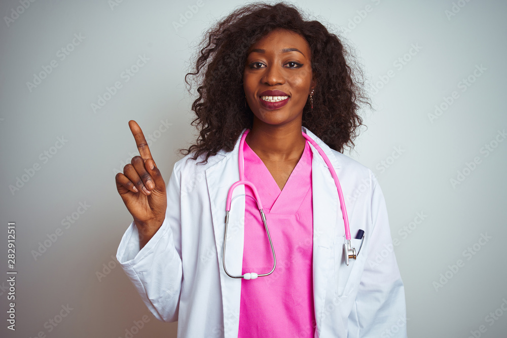 African american doctor woman wearing pink stethoscope over isolated white background showing and pointing up with finger number one while smiling confident and happy. African american doctor woman wearing pink stethoscope over isolated white background showing and pointing up with finger number one while smiling confident and happy.