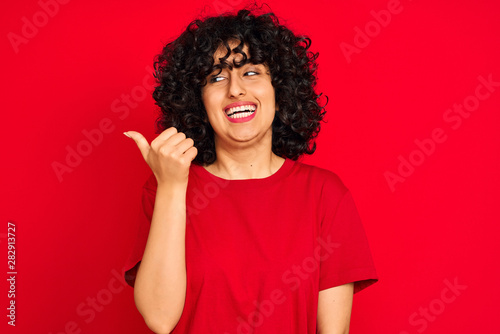 Young arab woman with curly hair wearing casual t-shirt over isolated red background smiling with happy face looking and pointing to the side with thumb up.