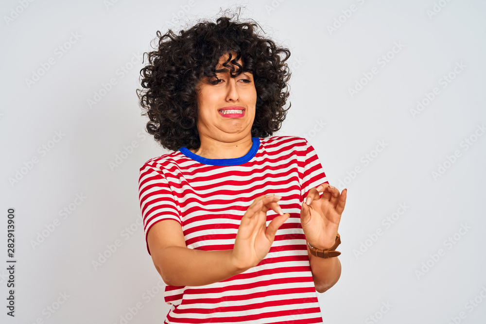 Fototapeta premium Young arab woman with curly hair wearing striped t-shirt over isolated white background disgusted expression, displeased and fearful doing disgust face because aversion reaction. With hands raised.