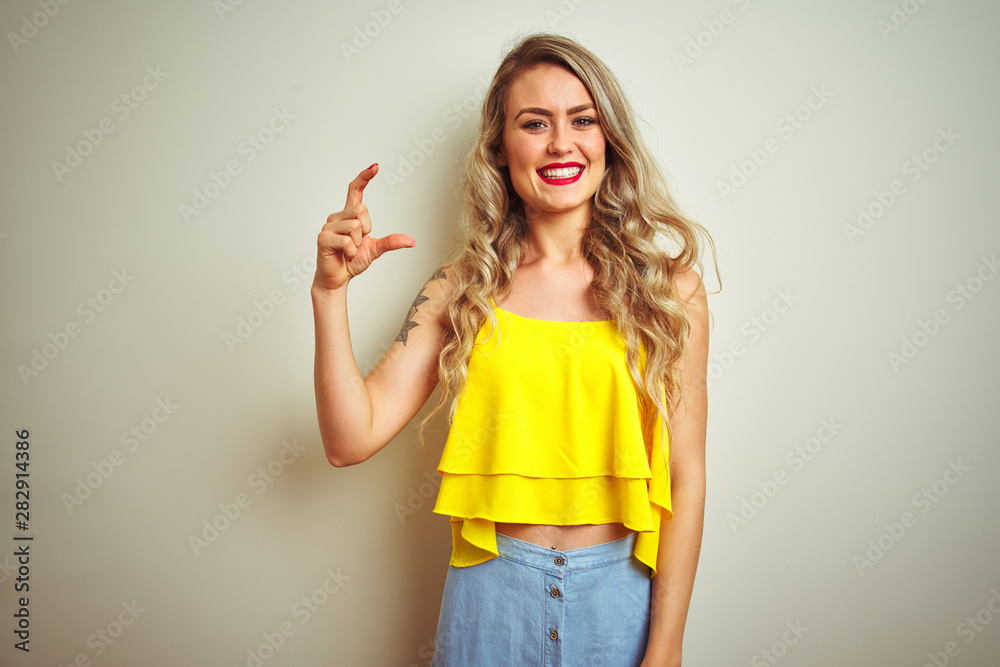 Young beautiful woman wearing yellow t-shirt standing over white isolated background smiling and confident gesturing with hand doing small size sign with fingers looking and the camera. Young beautiful woman wearing yellow t-shirt standing over white isolated background smiling and confident gesturing with hand doing small size sign with fingers looking and the camera.