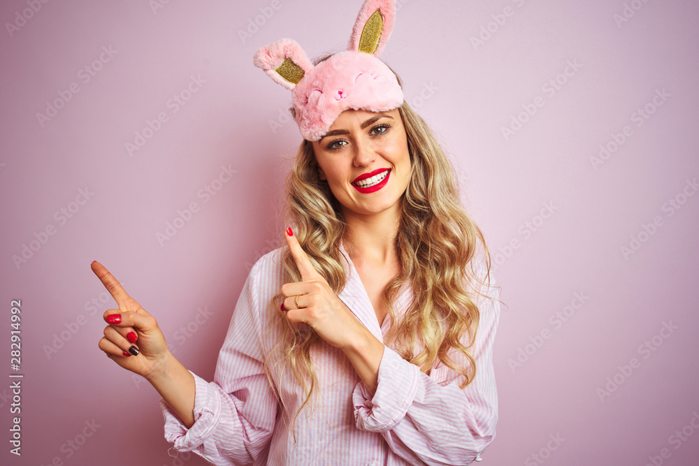 Young beautiful woman wearing pajama and sleep mask over pink isolated background smiling and looking at the camera pointing with two hands and fingers to the side. Young beautiful woman wearing pajama and sleep mask over pink isolated background smiling and looking at the camera pointing with two hands and fingers to the side.