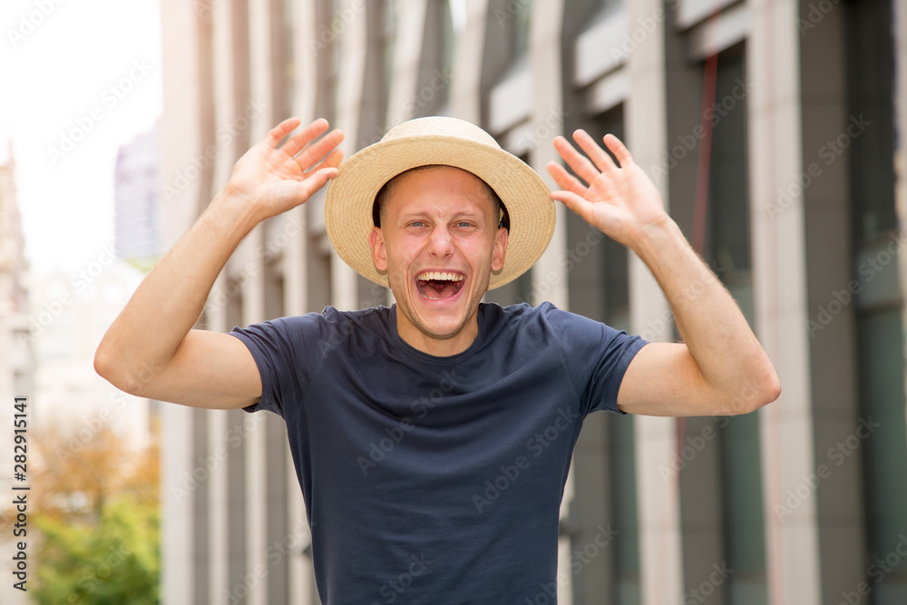 Greeting. Young happy man in a hat raised his hands up on the background of a European city.