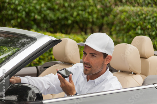 A man driving in his convertible car talks on speaker phone hold the phone close to his face. A businessman wears a business casual white baseball cap and a white long-sleeve button-down shirt.