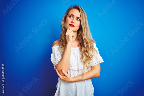 Young beautiful woman wearing white dress standing over blue isolated background with hand on chin thinking about question, pensive expression. Smiling with thoughtful face. Doubt concept.
