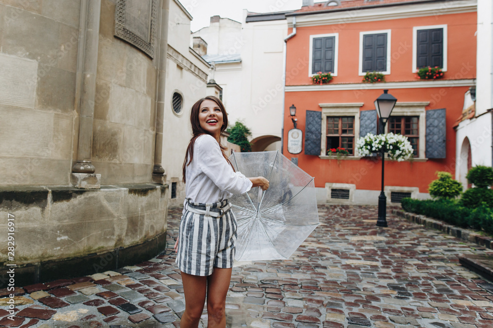 European girl walks around the city in summer, facial expression. Beautiful brunette young woman wearing shorts, walking on the street with transparent umbrella.