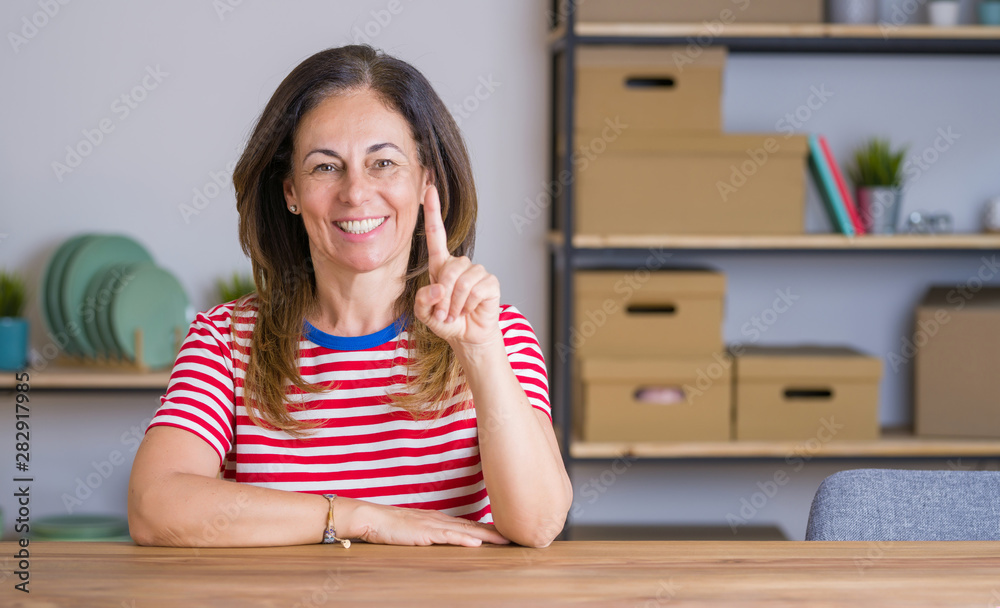 Middle age senior woman sitting at the table at home showing and pointing up with finger number one while smiling confident and happy. Middle age senior woman sitting at the table at home showing and pointing up with finger number one while smiling confident and happy.