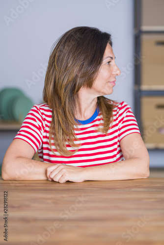 Middle age senior woman sitting at the table at home looking to side, relax profile pose with natural face with confident smile.