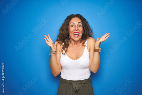 Middle age senior woman with curly hair standing over blue isolated background celebrating crazy and amazed for success with arms raised and open eyes screaming excited. Winner concept © Krakenimages.com