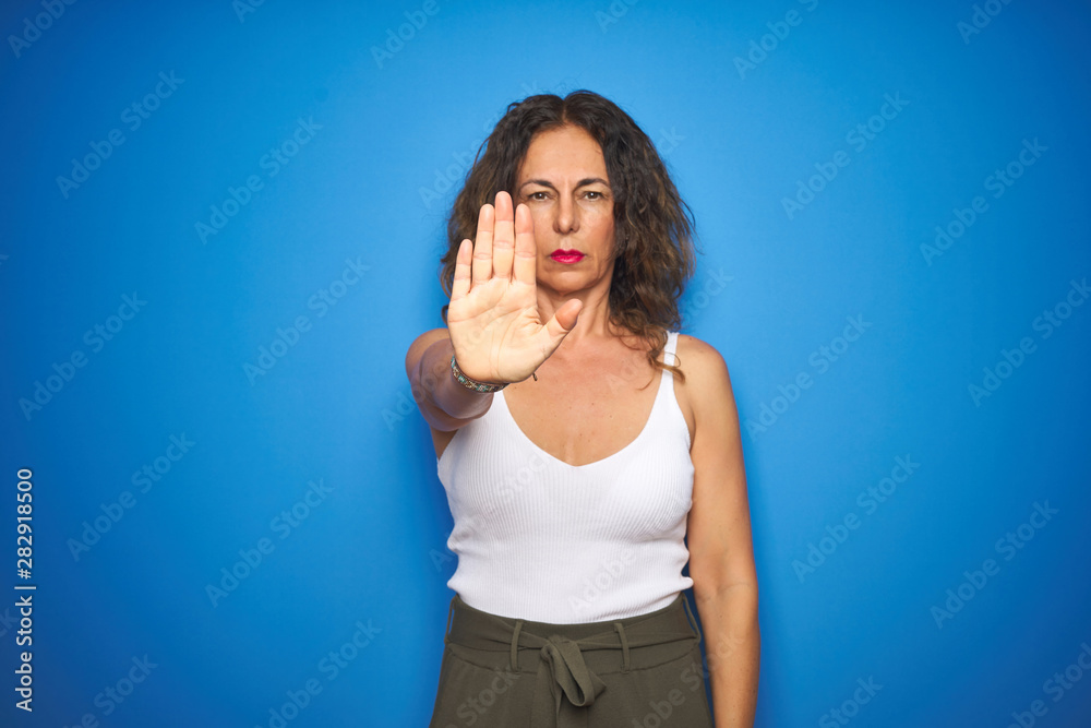 Middle age senior woman with curly hair standing over blue isolated background doing stop sing with palm of the hand. Warning expression with negative and serious gesture on the face. Middle age senior woman with curly hair standing over blue isolated background doing stop sing with palm of the hand. Warning expression with negative and serious gesture on the face.