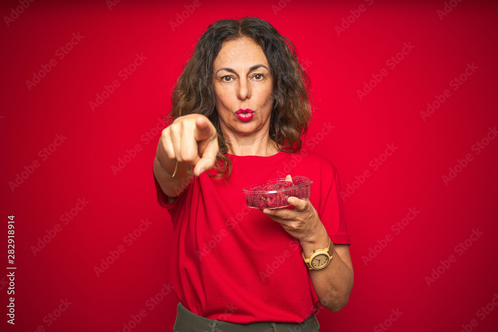 Middle age senior woman eating raspberries over red isolated background pointing with finger to the camera and to you, hand sign, positive and confident gesture from the front Middle age senior woman eating raspberries over red isolated background pointing with finger to the camera and to you, hand sign, positive and confident gesture from the front
