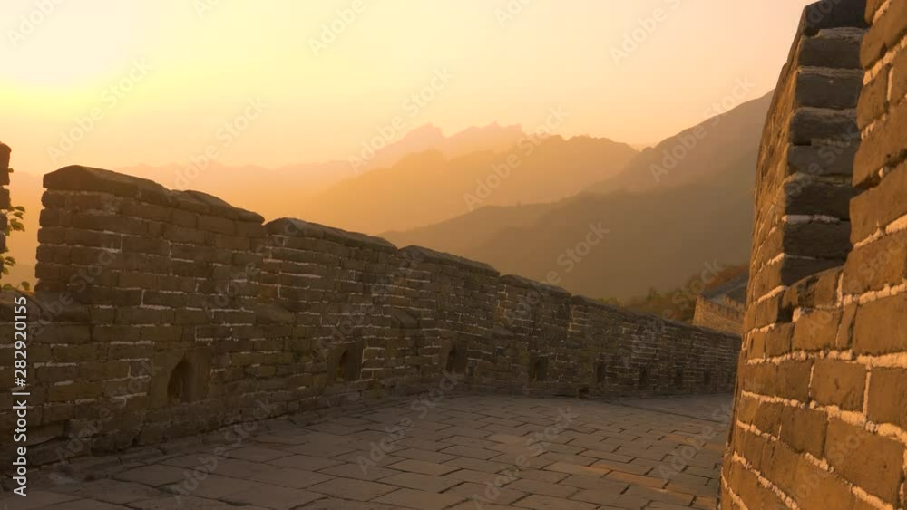 AERIAL: Beautiful burnt orange morning sunbeams shine on the majestic Great Wall of China. Autumn evening sky spans above the silhouettes of mountains in the distance and empty path on the Great Wall.