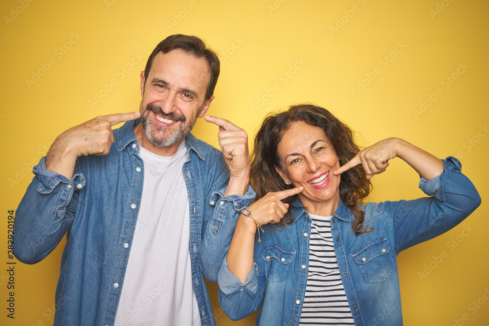 Beautiful middle age couple together wearing denim shirt over isolated yellow background smiling cheerful showing and pointing with fingers teeth and mouth. Dental health concept.