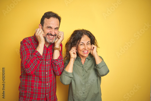 Beautiful middle age couple over isolated yellow background covering ears with fingers with annoyed expression for the noise of loud music. Deaf concept.