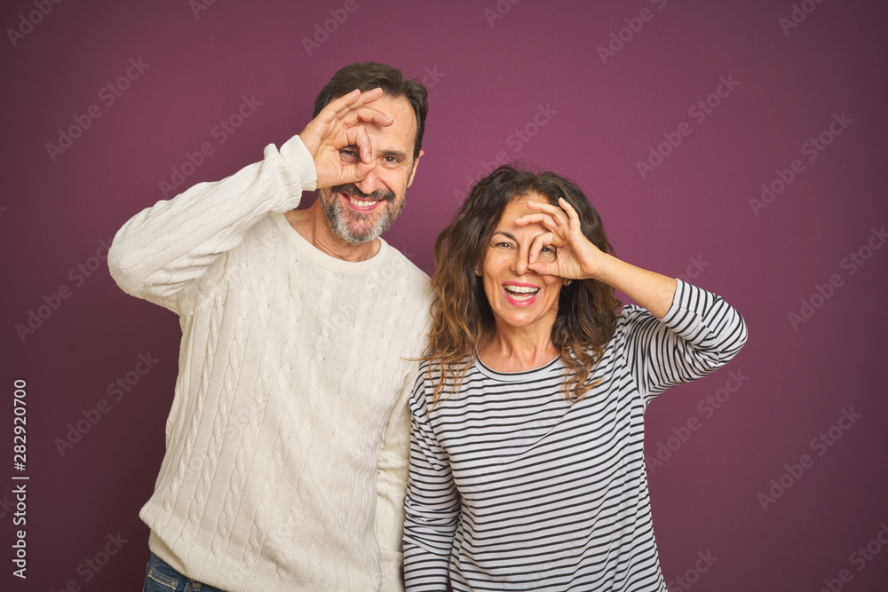Naklejka premium Beautiful middle age couple wearing winter sweater over isolated purple background doing ok gesture with hand smiling, eye looking through fingers with happy face.