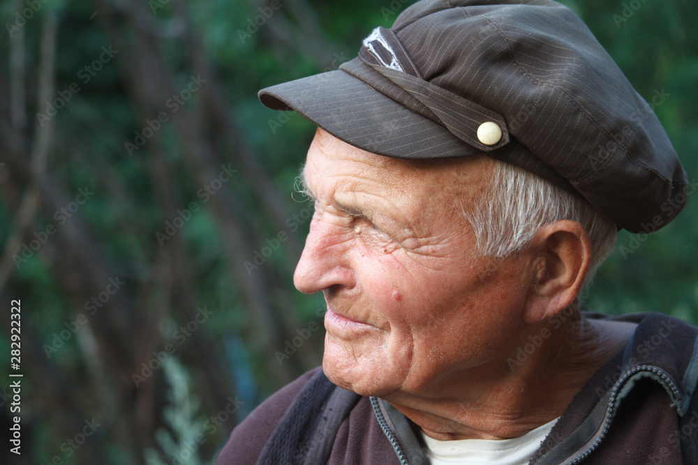 Close-up portrait of a very old man in a cap on a blurred background of green trees, selective focus