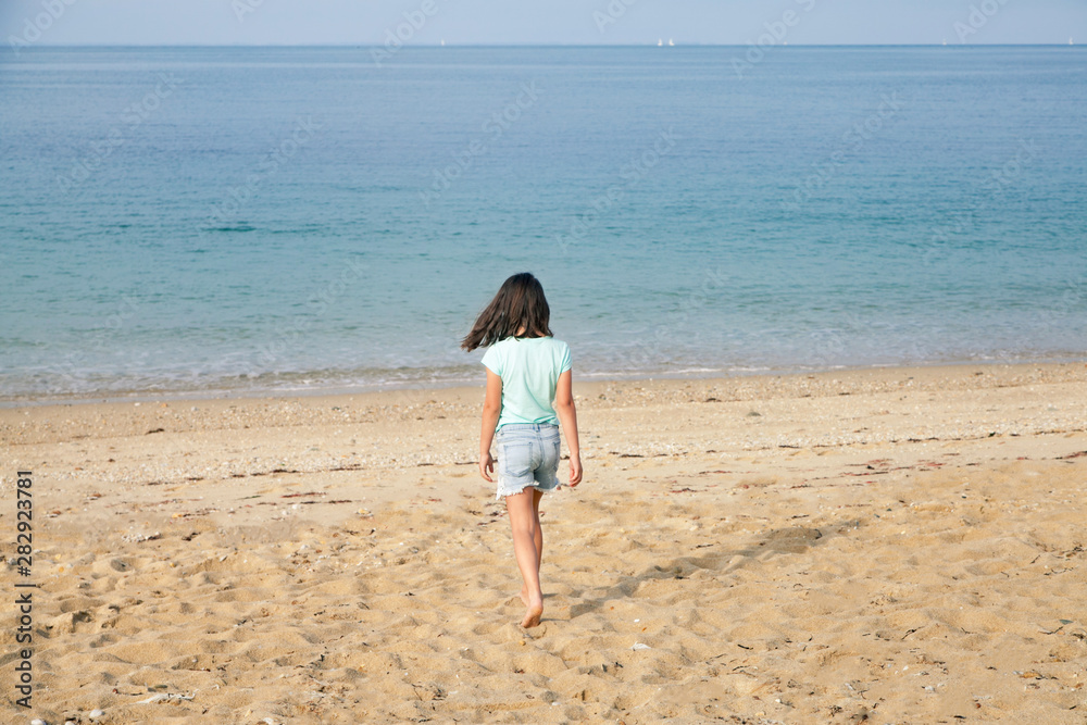 Young girl going away on sea background. Happy child in the ocean. White sand on the beach.