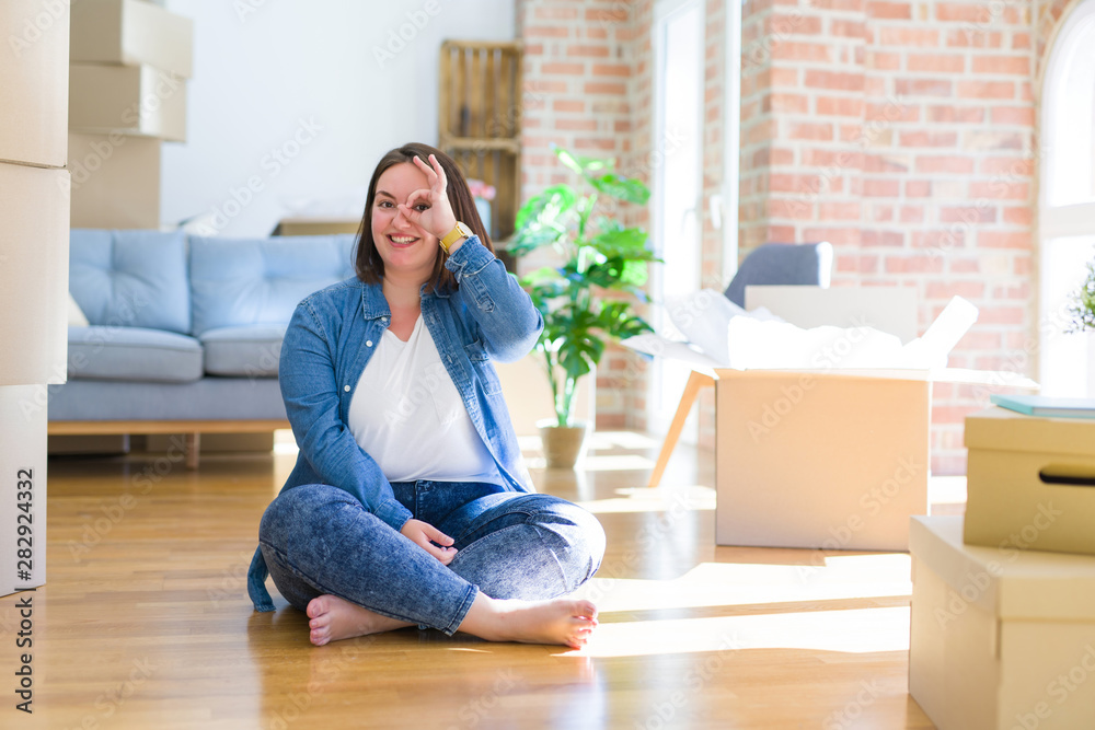 Young plus size woman sitting on the floor around cardboard boxes moving to a new home doing ok gesture with hand smiling, eye looking through fingers with happy face.