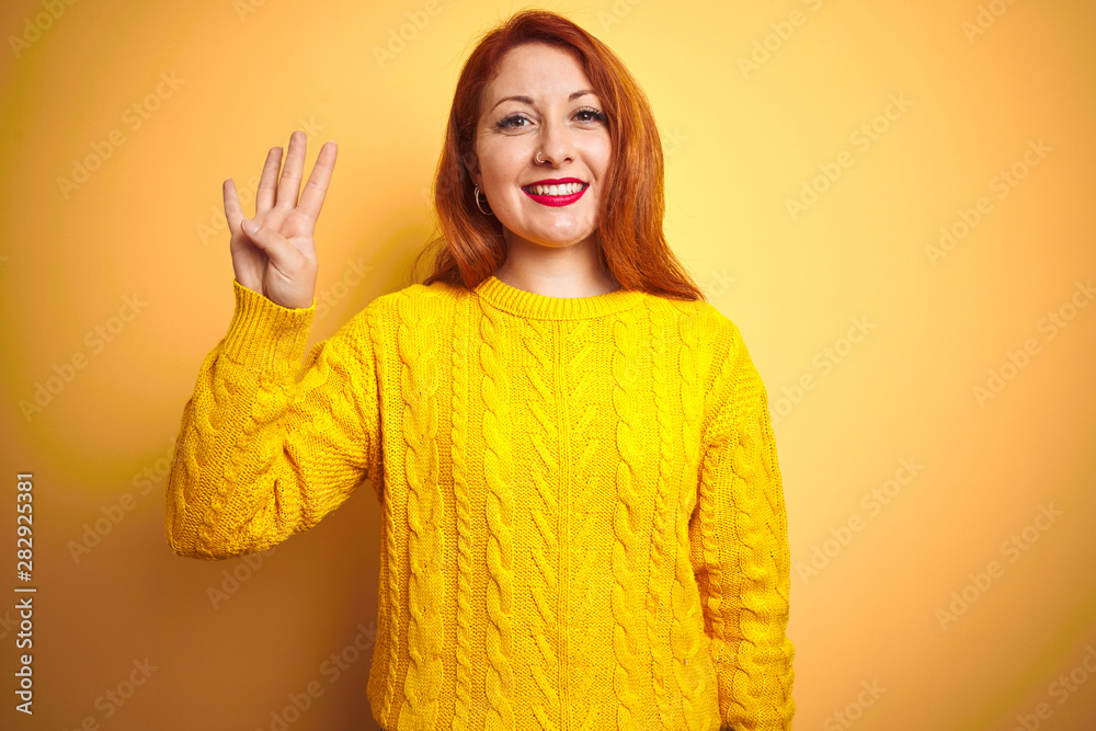 Beautiful redhead woman wearing winter sweater standing over isolated yellow background showing and pointing up with fingers number four while smiling confident and happy. Beautiful redhead woman wearing winter sweater standing over isolated yellow background showing and pointing up with fingers number four while smiling confident and happy.