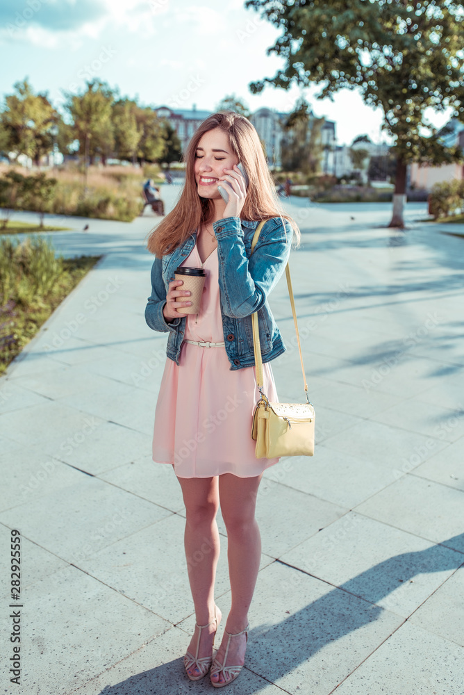 beautiful girl in dress and jeans, in summer in city, stands her hand phone, rings smartphone, application, online social networks, happy laughs, smiles, joyful cheerful, coffee tea in a cup beautiful girl in dress and jeans, in summer in city, stands her hand phone, rings smartphone, application, online social networks, happy laughs, smiles, joyful cheerful, coffee tea in a cup