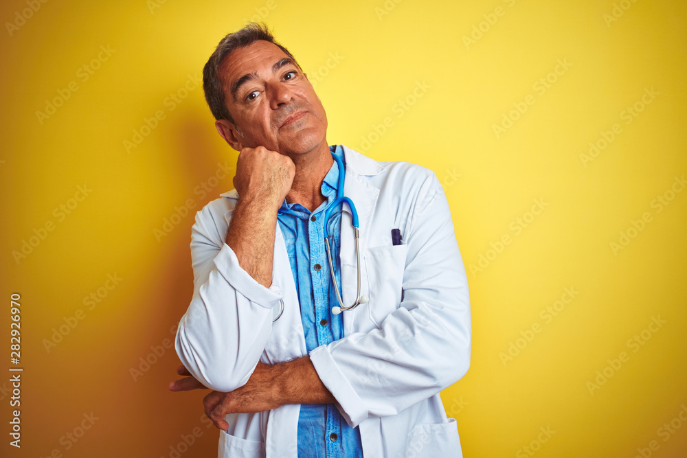 Handsome middle age doctor man wearing stethoscope over isolated yellow background with hand on chin thinking about question, pensive expression. Smiling with thoughtful face. Doubt concept.