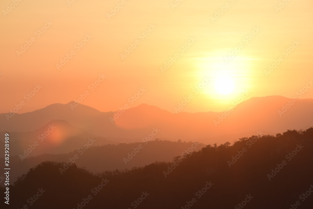 Picture of a sun setting behind a dense forest area followed by mountains.	