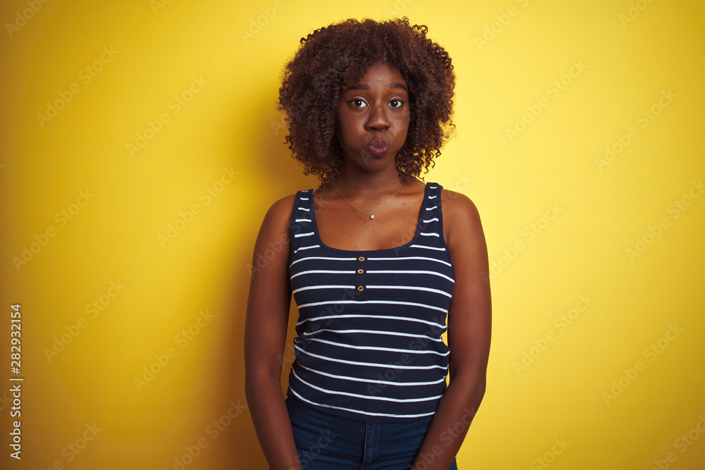 Young african afro woman wearing striped t-shirt over isolated yellow background puffing cheeks with funny face. Mouth inflated with air, crazy expression. Young african afro woman wearing striped t-shirt over isolated yellow background puffing cheeks with funny face. Mouth inflated with air, crazy expression.