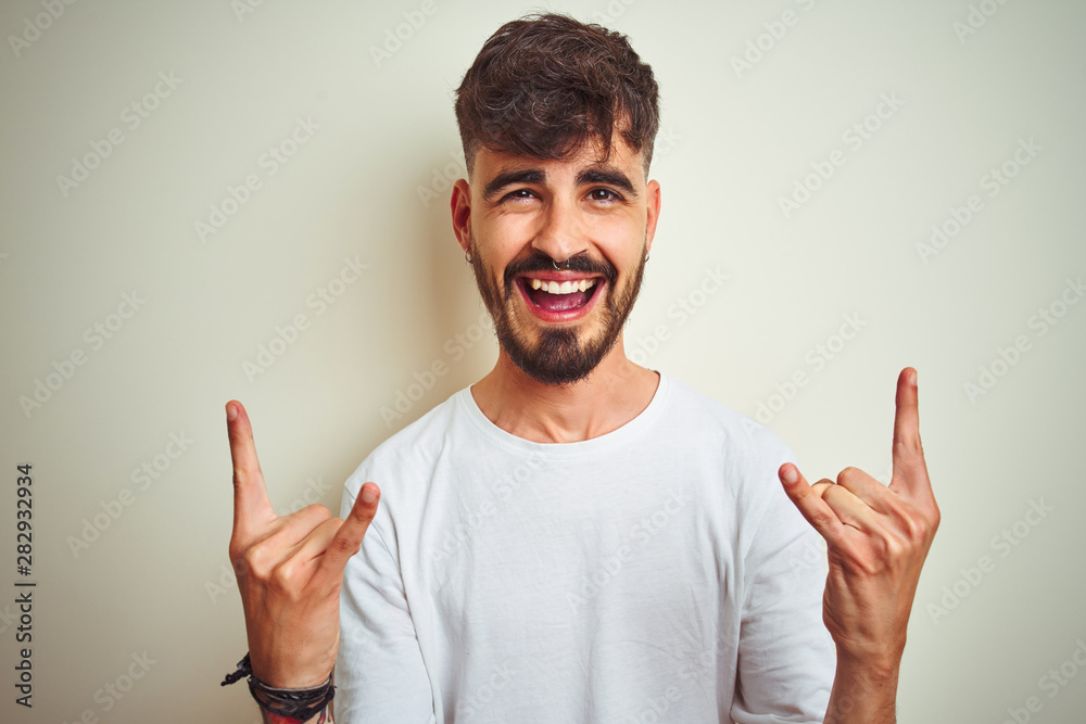 Young man with tattoo wearing t-shirt standing over isolated white background shouting with crazy expression doing rock symbol with hands up. Music star. Heavy concept. Young man with tattoo wearing t-shirt standing over isolated white background shouting with crazy expression doing rock symbol with hands up. Music star. Heavy concept.