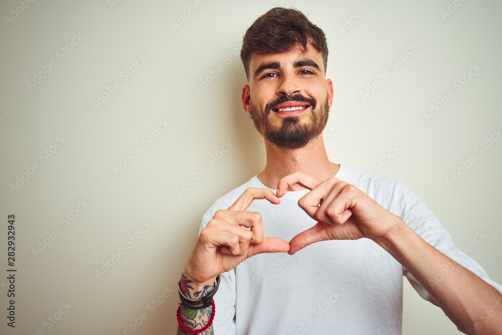 Young man with tattoo wearing t-shirt standing over isolated white background smiling in love doing heart symbol shape with hands. Romantic concept. Young man with tattoo wearing t-shirt standing over isolated white background smiling in love doing heart symbol shape with hands. Romantic concept.
