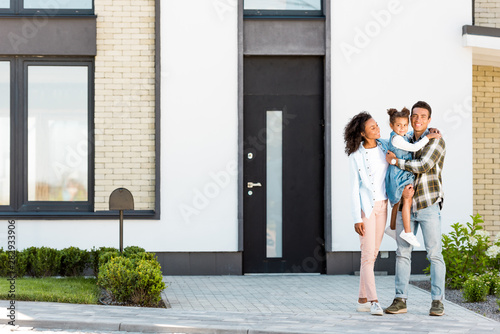 full length view of african american family standing near new house while father holding kid and looking at camera