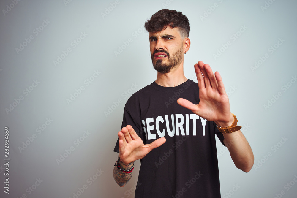 Young safeguard man with tattoo wering security uniform over isolated white background afraid and terrified with fear expression stop gesture with hands, shouting in shock. Panic concept. Young safeguard man with tattoo wering security uniform over isolated white background afraid and terrified with fear expression stop gesture with hands, shouting in shock. Panic concept.
